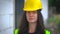 Young woman in yellow hard hat and green high visibility vest, long dark hair, looking to camera, smiling at the end. Blurred