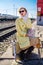 A young woman in a yellow raincoat and dark glasses sits on a suitcase on the platform of a railway station