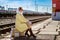 A young woman in a yellow raincoat and dark glasses sits on a suitcase on the platform of a railway station
