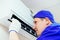 A young worker in a blue baseball cap and gloves inspects and maintains the air conditioner in the room.