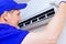 A young worker in a blue baseball cap and gloves inspects and maintains the air conditioner in the room.