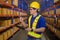 Young worker wearing helmet checking inventory and counting product on shelf in modern warehouse