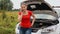 Young worried woman standing next to broken car in field and calling service