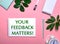 YOUR FEEDBACK MATTERS is written in green on a white notepad on a pink background surrounded by notepads, pens, white alarm clock