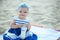 Ð¡ute baby girl in nice striped dress and blue headband sitting on the sand and trying to open a bottle with childâ€™s milk