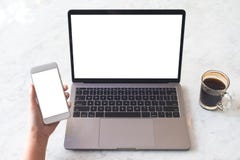 A Hand Holding White Mobile Phone With Blank Screen With Laptop With Blank White Desktop Screen And Coffee Cup On Table Stock Photography