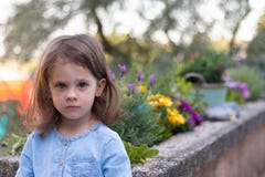 A Little Beautiful Preschooler Girl In A Denim Dress Stands On A Blurred Background Near Bright Flowers Stock Photography
