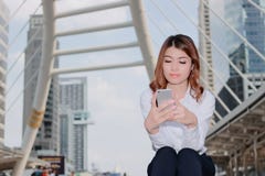 Aantrekkelijke jonge Aziatische vrouw die in wit overhemd mobiele smartphone in haar handen stedelijke de bouwachtergrond bekijkt stock afbeeldingen Aantrekkelijke jonge Aziatische vrouw die in wit overhemd mobiele smartphone in haar handen stedelijke de bouwachtergrond bekijkt stock afbeeldingen