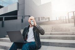 Aantrekkelijke volwassen succesvolle kale gebaarde mens in zwart jasje gebruikend laptop en drinkend koffie in treden bij stad stock foto Aantrekkelijke volwassen succesvolle kale gebaarde mens in zwart jasje gebruikend laptop en drinkend koffie in treden bij stad stock foto