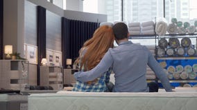 Achtermening die van een paar wordt geschoten die, zittend op een nieuw bed bij meubilairopslag koesteren stock footage Achtermening die van een paar wordt geschoten die, zittend op een nieuw bed bij meubilairopslag koesteren stock footage