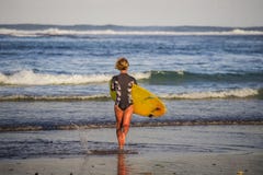 Achtermening van jong aantrekkelijk en sportief surfermeisje in koel zwempak bij de raad van de strand dragende branding in het o royalty-vrije stock foto Achtermening van jong aantrekkelijk en sportief surfermeisje in koel zwempak bij de raad van de strand dragende branding in het o royalty-vrije stock foto