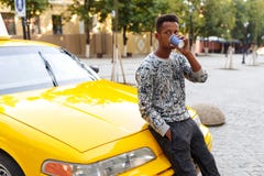 Afrikanischer Mann, der einen Kaffee gesetzt auf der Haube eines Taxis, schauend zu einer Seite, auf einem Stra?enhintergrund tri lizenzfreie stockfotos Afrikanischer Mann, der einen Kaffee gesetzt auf der Haube eines Taxis, schauend zu einer Seite, auf einem Stra?enhintergrund tri lizenzfreie stockfotos