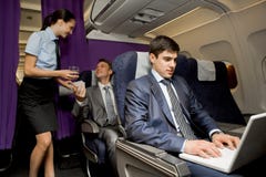 In airplane. Image of busy male typing on laptop with pretty stewardess giving glass of water to successful businessman on background In airplane. Image of busy male typing on laptop with pretty stewardess giving glass of water to successful businessman on background
