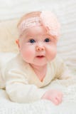 Amazed baby girl with chubby cheeks and big blue eyes wearing white clothes and pink band with flower lying on bed royalty free stock image Amazed baby girl with chubby cheeks and big blue eyes wearing white clothes and pink band with flower lying on bed royalty free stock image