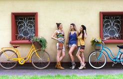 As meninas chiques do boho feliz levantam com as bicicletas perto da fachada da casa fotografia de stock As meninas chiques do boho feliz levantam com as bicicletas perto da fachada da casa fotografia de stock