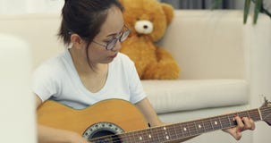 Asian young girl sitting on long sofa chair concentrating focused learning to play guitar at home.