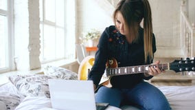 Attractive young girl learning to play electric guitar with notebook sit on bed in bedroom at home