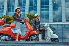 Attraktiva romantiska par, en stilig man och sexig kvinnlig som sitter på retro italienska sparkcyklar mot en skyskrapa royaltyfria bilder Attraktiva romantiska par, en stilig man och sexig kvinnlig som sitter på retro italienska sparkcyklar mot en skyskrapa royaltyfria bilder