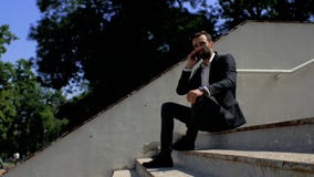 Attraktiver Geschäftsmann des jungen Mannes mit einem Bart, der in einem Park auf einem konkreten Treppenhaus sitzt und am Telefo stock video Attraktiver Geschäftsmann des jungen Mannes mit einem Bart, der in einem Park auf einem konkreten Treppenhaus sitzt und am Telefo stock video