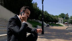 Attraktiver Geschäftsmann des jungen Mannes mit einem Bart, der in einem Park auf einem konkreten Treppenhaus sitzt und am Telefo stock footage Attraktiver Geschäftsmann des jungen Mannes mit einem Bart, der in einem Park auf einem konkreten Treppenhaus sitzt und am Telefo stock footage