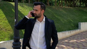 Attraktiver junger Geschäftsmann mit einem Bart in einem Hemd und in einer Jacke sprechend am Telefon im Park an einem klaren Som stock video Attraktiver junger Geschäftsmann mit einem Bart in einem Hemd und in einer Jacke sprechend am Telefon im Park an einem klaren Som stock video