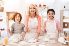 A avó feliz nova junto com netos felizes pequenos amassa a massa para cookies na cozinha foto de stock A avó feliz nova junto com netos felizes pequenos amassa a massa para cookies na cozinha foto de stock