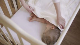 Baby girl 2 years old sleeping in a crib covered white blanket. Mom covers the baby with a blanket. Daytime sleep