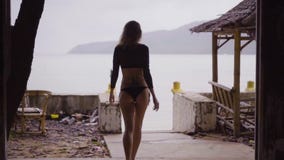 Back view of blonde woman wearing a black elegant swimsuit standing in terrace window of abandoned building over sea.