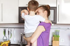Back view of young slim mother keeps son on hands, stand at kitchen against modern furniture, going to eat supper. Busy cute woman stock photos Back view of young slim mother keeps son on hands, stand at kitchen against modern furniture, going to eat supper. Busy cute woman stock photos