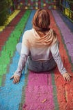 Young woman wearing casual clothes sits back to camera on rainbow painted stairs