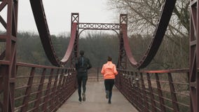 Backside view of young sportive couple running across bridge in parkland. Woman and man athletes in bright sportswear jogging outd
