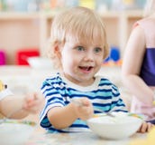 Bambino sorridente divertente che mangia dal piatto nell'asilo fotografia stock Bambino sorridente divertente che mangia dal piatto nell'asilo fotografia stock