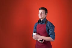 Barman de sourire beau avec les cheveux fonc?s dans le tablier de Bourgogne tenant la tasse de papier et le smartphone image libre de droits Barman de sourire beau avec les cheveux fonc?s dans le tablier de Bourgogne tenant la tasse de papier et le smartphone image libre de droits
