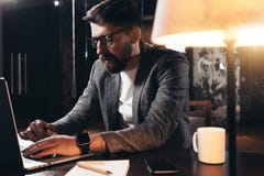 Bearded young businessman working in loft space at night. Coworker sits by the wooden table with lamp and office tools royalty free stock photo Bearded young businessman working in loft space at night. Coworker sits by the wooden table with lamp and office tools royalty free stock photo
