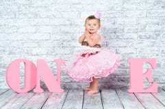 Beautiful And Cute Girl In A Pink Dress With The Letters One On Her First Birthday. Emotional Girl. Stock Photo