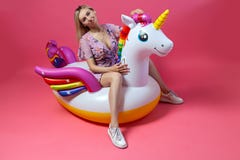 A beautiful blonde girl in a sexy sundress with slim legs in white sneakers sits on an inflatable multi-colored unicorn royalty free stock images A beautiful blonde girl in a sexy sundress with slim legs in white sneakers sits on an inflatable multi-colored unicorn royalty free stock images