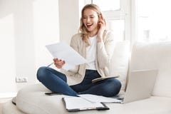 Beautiful blonde woman posing sitting indoors at home using laptop computer talking by mobile phone stock image Beautiful blonde woman posing sitting indoors at home using laptop computer talking by mobile phone stock image