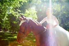 A beautiful bride in a white dress riding a horse in a green sunny summer park