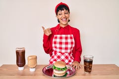Beautiful brunettte woman cooking tasty hamburguer pointing thumb up to the side smiling happy with open mouth