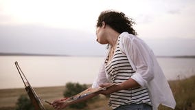 Beautiful curly woman artist concentrated on painting process. Working on future picture, using a paintbrush applies oil stock video Beautiful curly woman artist concentrated on painting process. Working on future picture, using a paintbrush applies oil stock video