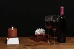 Beautiful etched wine glasses and bottle of red wine, red candle, wrapped present with bow on wooden table with name tag on dark royalty free stock photography Beautiful etched wine glasses and bottle of red wine, red candle, wrapped present with bow on wooden table with name tag on dark royalty free stock photography