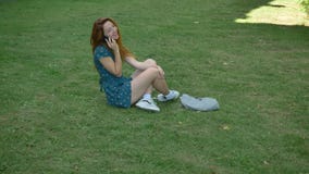 Beautiful ginger young woman talking on phone and laughing, sitting on grass, happy and cheerful female speaking stock video footage Beautiful ginger young woman talking on phone and laughing, sitting on grass, happy and cheerful female speaking stock video footage