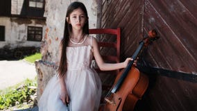 Beautiful girl in a pink dress sits on a red chair with a cello