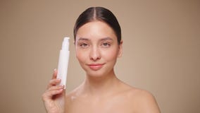 Beautiful woman with bottle of moisturizer cream. Video of woman with perfect skin on beige background. Beauty and Skin care concept. Slow motion