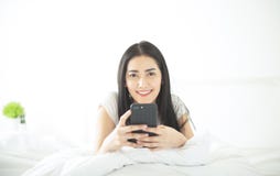 Beautiful young Asian smiling brunette woman lying in white bed and using a mobile in her bedroom and looking at camera. Relax and stock photography Beautiful young Asian smiling brunette woman lying in white bed and using a mobile in her bedroom and looking at camera. Relax and stock photography