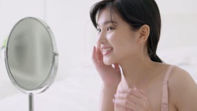 Beautiful young asian woman smiling look at mirror of checking face with skin care and cosmetic for rejuvenation