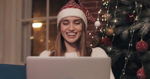 Beautiful young blond caucasian girl wearing santas hat sitting near Christmas tree using her laptop chatting making stock footage Beautiful young blond caucasian girl wearing santas hat sitting near Christmas tree using her laptop chatting making stock footage