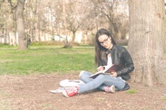 Beautiful young school or college girl with long hair, eyeglasses and black leather jacket sitting on the ground in the park readi royalty free stock photo Beautiful young school or college girl with long hair, eyeglasses and black leather jacket sitting on the ground in the park readi royalty free stock photo