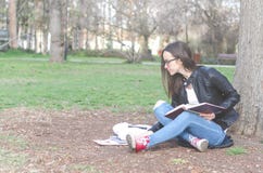 Beautiful young school or college girl with long hair, eyeglasses and black leather jacket sitting on the ground in the park readi royalty free stock images Beautiful young school or college girl with long hair, eyeglasses and black leather jacket sitting on the ground in the park readi royalty free stock images