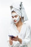 Beautiful young woman dressed in white clothes with a white towel on her hair and cosmetic mask on her face holds mobile royalty free stock image Beautiful young woman dressed in white clothes with a white towel on her hair and cosmetic mask on her face holds mobile royalty free stock image